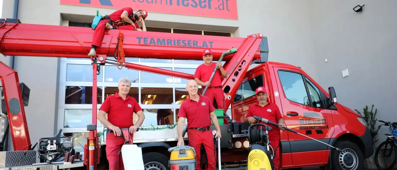 Sechs Mitarbeiter in roter Arbeitskleidung vor einem roten Firmentransporter mit Hubsteiger; einer liegt oben auf dem Ausleger, vor der Gruppe stehen Einscheibenmaschine, Nasssauger und Hochdruckreiniger.
