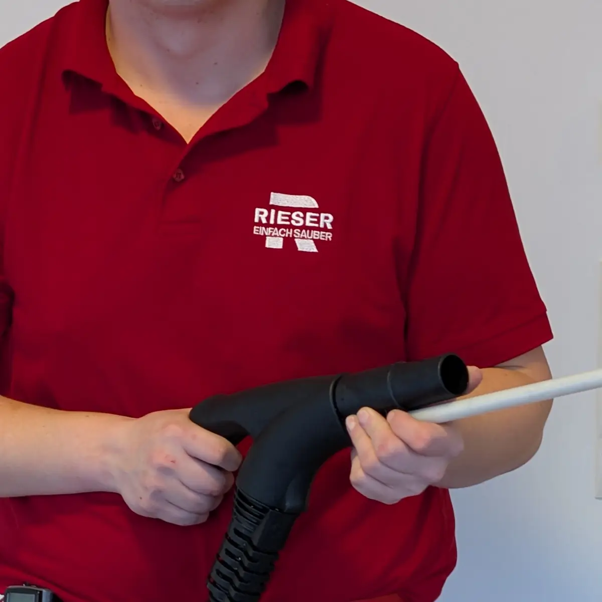 Nahaufnahme eines roten Poloshirts mit dem gestickten Schriftzug „RIESER – einfach sauber“. Darunter halten zwei Hände Griff und Rohr einer Dampfreiniger-Lanze.