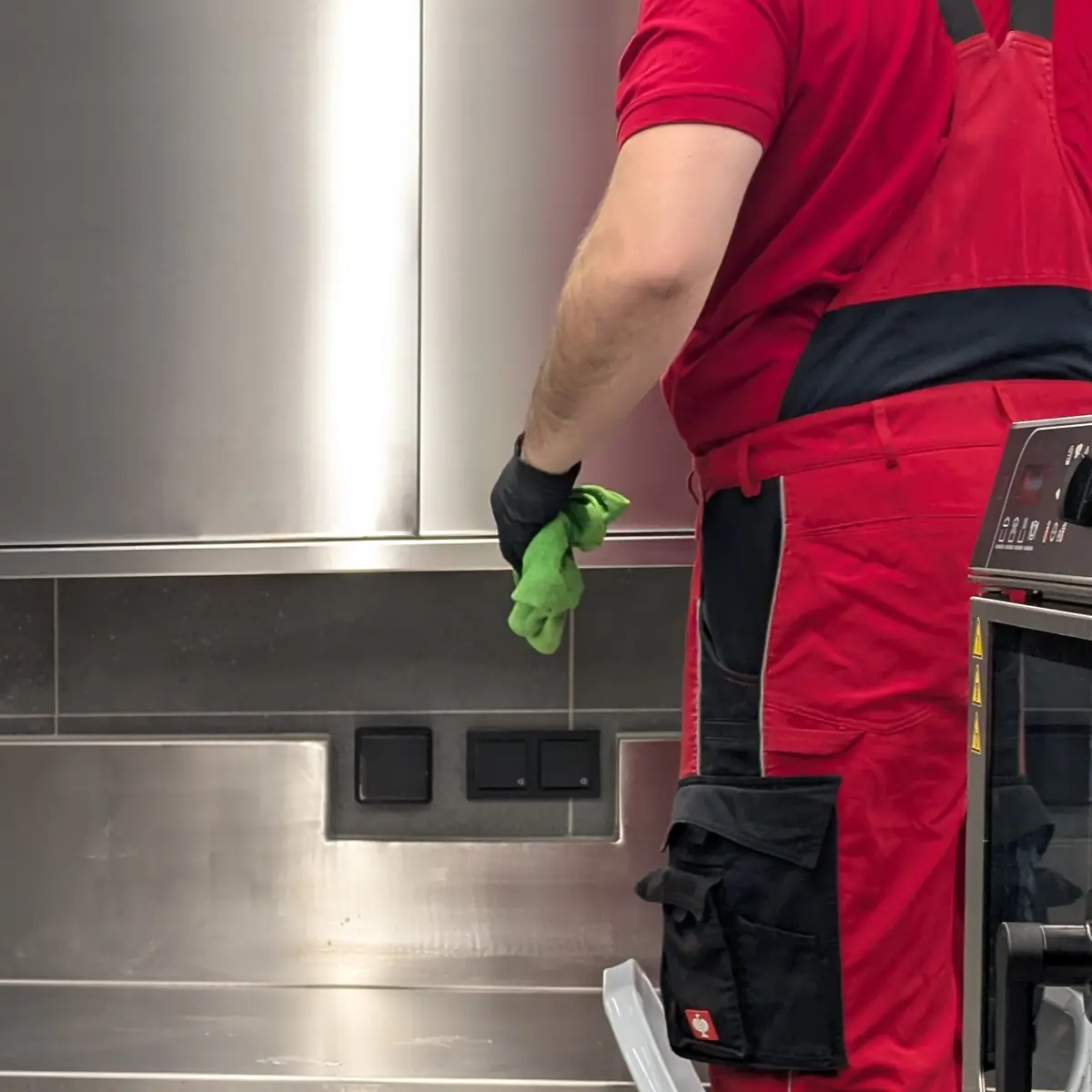 Nahaufnahme: eine Hand im schwarzen Handschuh hält ein grünes Mikrofasertuch vor einer Edelstahlwand in einer Großküche.