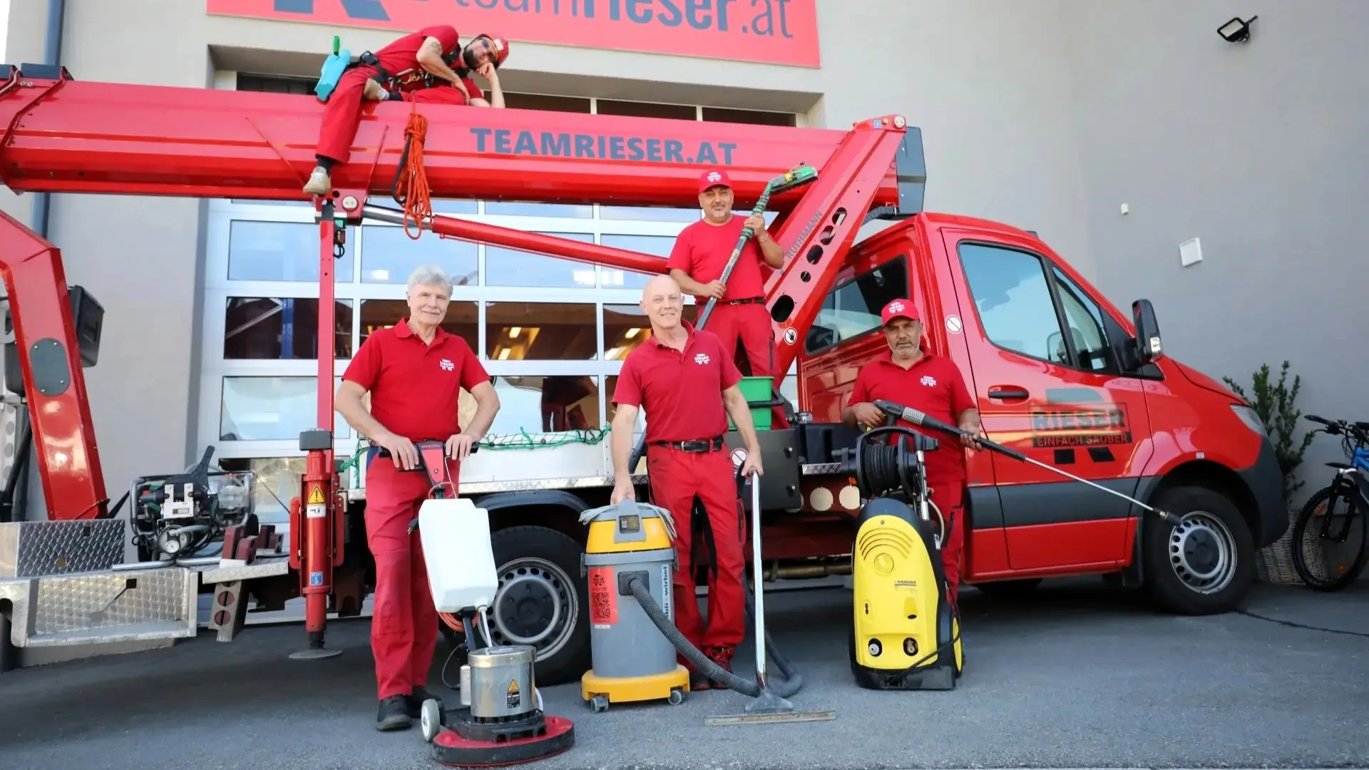 Fünf Mitarbeiter von Team Rieser in roter Arbeitskleidung stehen vor einem roten Firmentransporter mit Hubsteiger. Vor ihnen stehen Einscheibenmaschine, Nasssauger und Hochdruckreiniger.