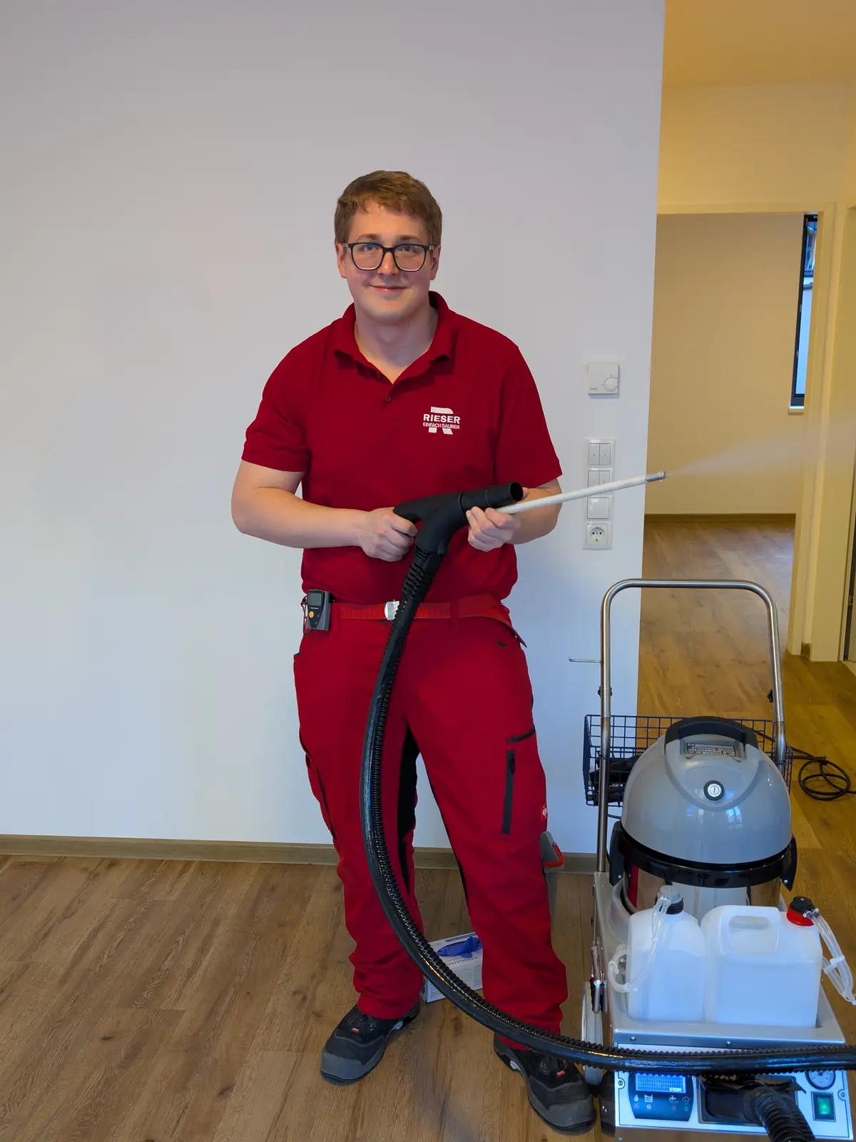 Ein junger Mitarbeiter in roter Arbeitskleidung steht mit der Lanze eines Dampfreinigers in einer leeren Wohnung; rechts neben ihm steht das Gerät mit Kanistern auf einem Wagen.