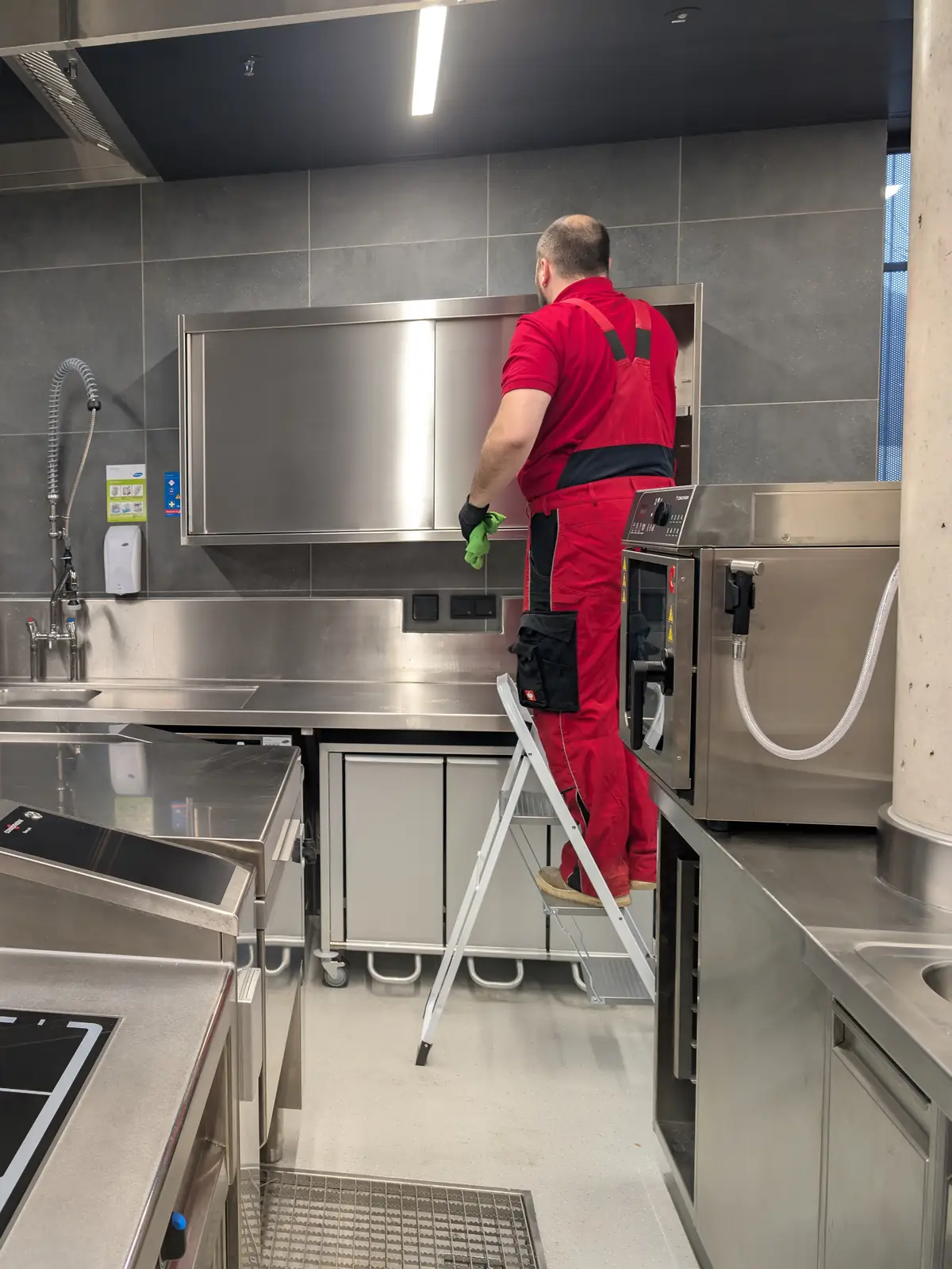 Ein Mitarbeiter steht auf einer Trittleiter und wischt in einer Großküche einen Edelstahl-Hängeschrank ab.
