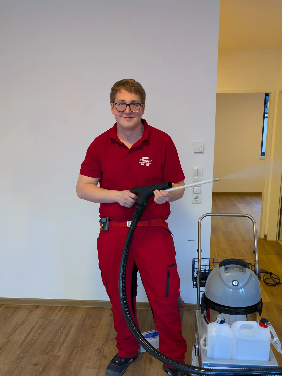 Ein junger Mitarbeiter in roter Arbeitskleidung steht mit der Lanze eines Dampfreinigers in einer leeren Wohnung und schaut lächelnd in die Kamera.