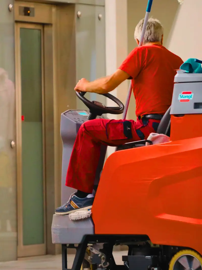 Ein Mitarbeiter fährt eine orange Aufsitz-Scheuersaugmaschine durch eine Eingangshalle.