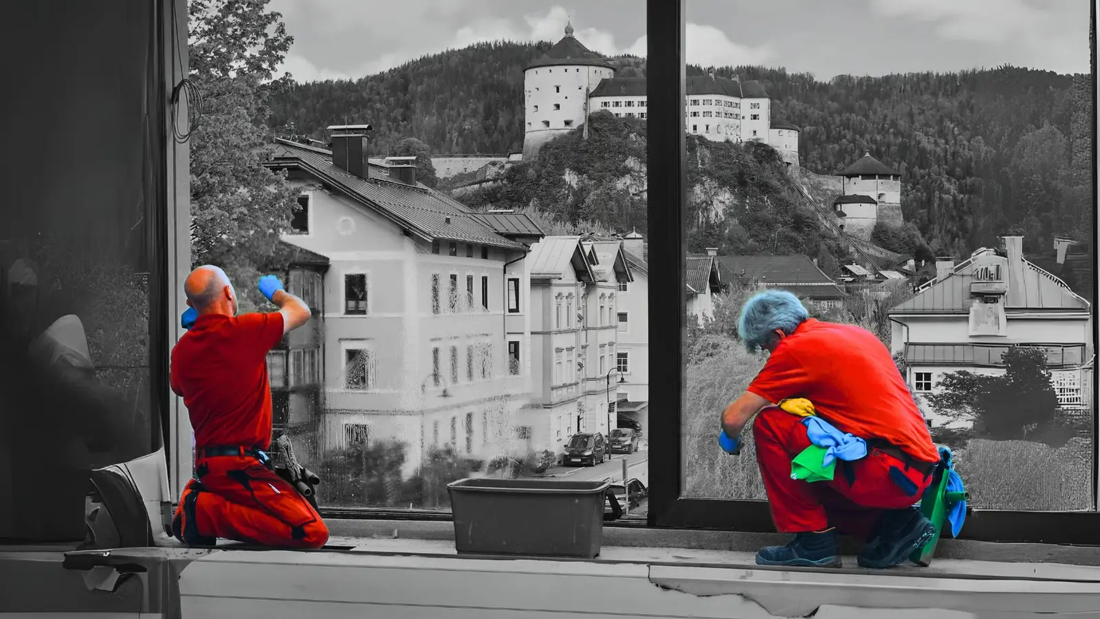 Zwei Mitarbeiter reinigen von innen ein großes Fenster. Dahinter liegt die Festung Kufstein über den Dächern der Stadt.