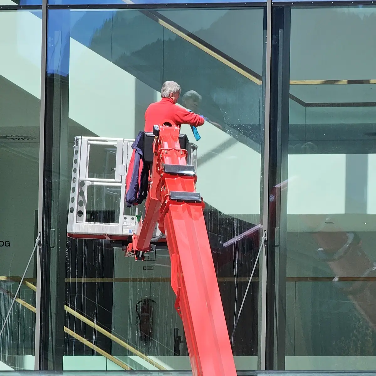 Ein Glasreiniger arbeitet vom Korb eines roten Hubsteigers aus an einer großen Glasfassade.