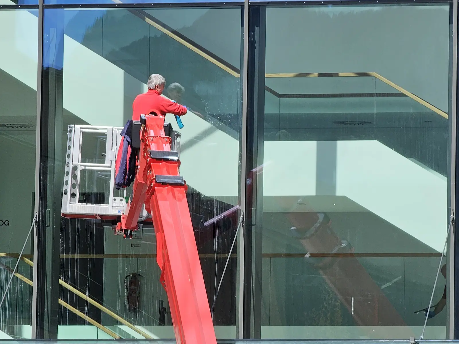 Ein Glasreiniger arbeitet vom Korb eines roten Hubsteigers aus an einer großen Glasfassade.