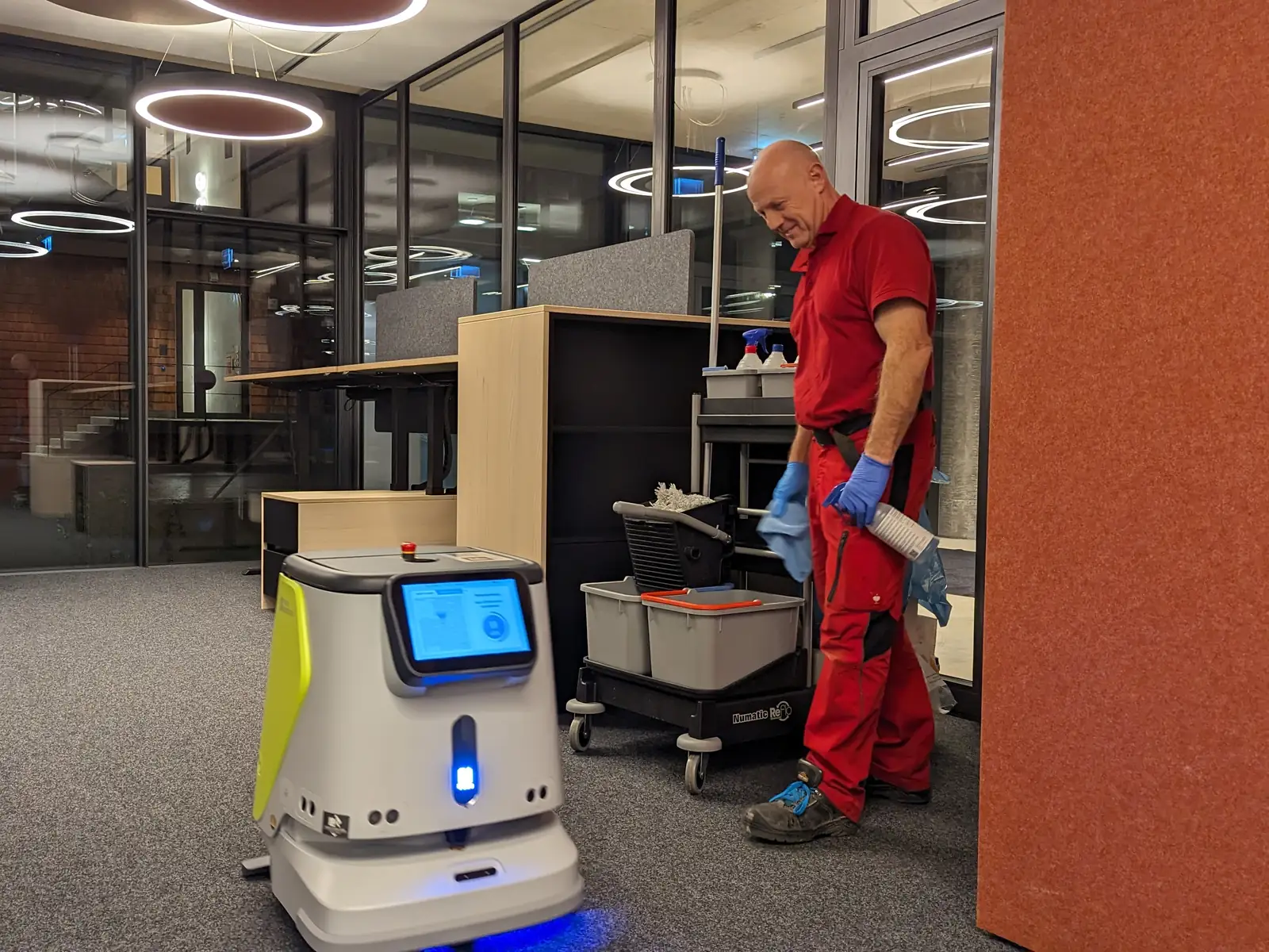 Ein Mitarbeiter mit Reinigungswagen und ein Reinigungsroboter arbeiten in einem Großraumbüro.