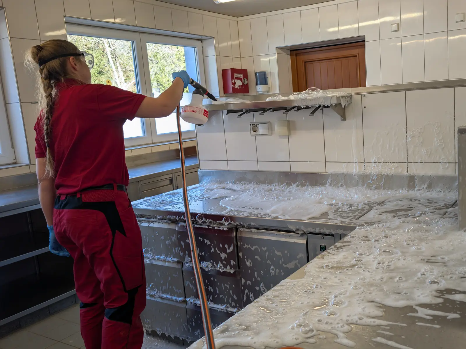 Eine Mitarbeiterin sprüht mit einer Sprühlanze Reinigungsschaum auf die Edelstahlflächen einer Bäckerei.