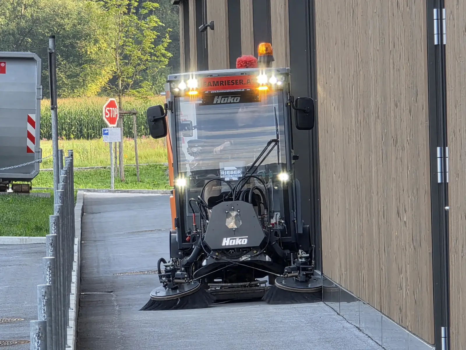 Eine Aufsitz-Kehrmaschine mit Rundumleuchte reinigt am frühen Morgen die Zufahrt neben einer Halle.